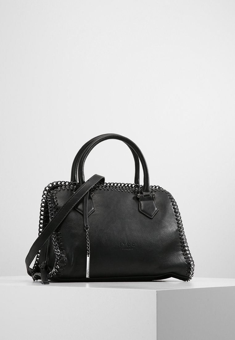 Sac à main en cuir noir avec garniture chaîne sur les bords, deux poignées et une bandoulière amovible, présenté sur un socle blanc sur fond gris.