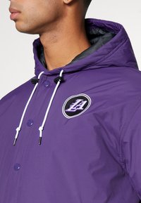 Lila huva jacka med glänsande yta, utrustad med vita snoddar, framknappar och en cirkulär broderad logotyp i svart och lila.