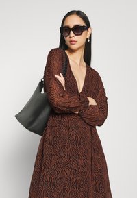 Vestido castanho de mangas longas com um padrão arrojado de zebras, combinado com uma bolsa preta em forma de balde. A modelo usa óculos de sol oversized escuros e brincos.