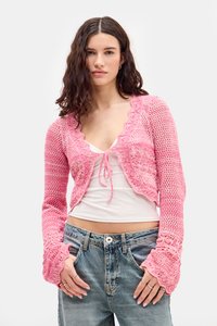 Cárdigan de ganchillo en rosa brillante, con un escote ondulado y mangas acampanadas, llevado sobre una blusa blanca y combinado con jeans azules.