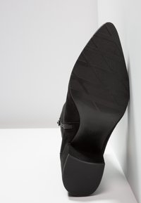 Semelle d'une bottine en daim noir montrant des motifs de grip texturés et un talon bloc, positionnée verticalement sur fond blanc.