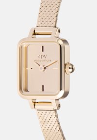 Goldfarbener rechteckiger Uhr mit glattem Ziffernblatt, minimalen Stundenmarkierungen und einem texturierten Mesh-Armband. Verfügt über eine auffällige Krone und ein Markenlogo.