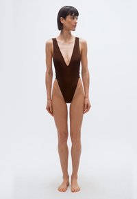 Γυναίκα όρθια με γυμνά πόδια, που φορεί καφέ μαγιό χωρίς μανίκια με βαθύ V λαιμόκοψη, κοιτάζει προς τα αριστερά της σε ένα απλό λευκό φόντο.