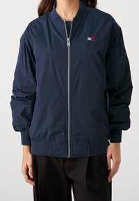 Tmavě modrá bomber bunda z lehké, hladké látky s předním zipem, žebrovanými manžetami a malým vyšitým logem na hrudi.