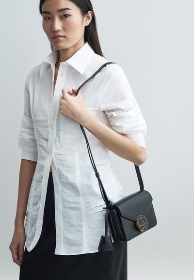 Hosszú sötét hajú nő fehér, ráncolt inget és fekete szoknyát visel, fekete bőr válltáskával a kezében, amelyen arany béke szimbólum csat található.