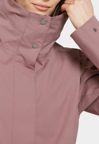 Ljusrosa vattentät jacka med hög krage, tryckknappar och slät textur; innehåller en närbild av krageområdet.