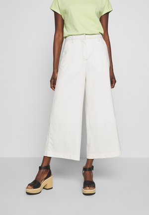 Mujer con pantalones blancos de talle alto y pierna ancha, camiseta de manga corta verde claro y sandalias negras de plataforma con tiras en el tobillo sobre un fondo gris.