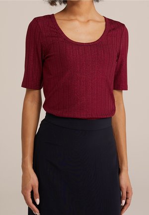 Kobieta ubrana w prążkowany bordowy top z krótkim rękawem i dekoltem w kształcie litery U, włożony do czarnej spódnicy z wysokim stanem, na jednolitym tle.