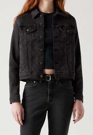 Femme portant une veste en jean noire sur un crop top noir ajusté, avec un jean noir taille haute et une ceinture noire avec boucle argentée.