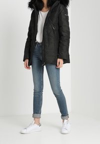 Svart pufferjacka med en huva kantad med päls, två fickor med dragkedja, ihop med ljusblå jeans och vita sneakers. Minimalistisk design.