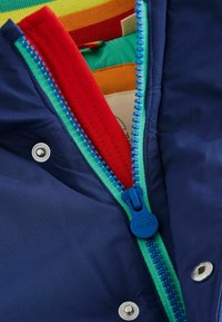 Marinejacke mit einem leuchtend blauen Reißverschluss, grünen Akzenten und rotem Futter. Mit silbernen Druckknöpfen und bunten Innenstofflagen.