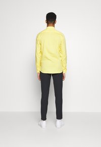 Geel overhemd met lange mouwen en knopenmanchetten, gecombineerd met donkere broeken en witte sneakers. Eenvoudig ontwerp, stof lijkt glad en licht van gewicht.