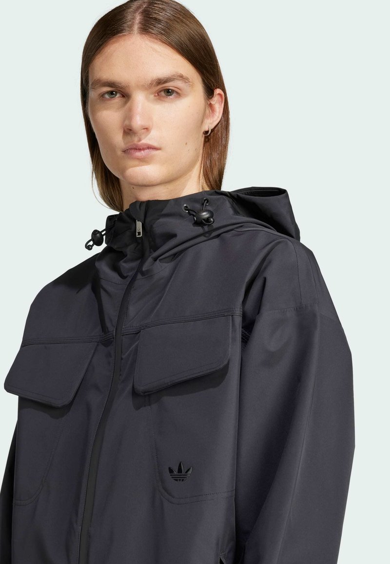 Veste noire imperméable avec fermeture éclair à l'avant, deux grandes poches poitrine, capuche ajustable et logo discret. Texture lisse, design décontracté.