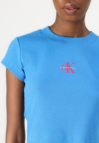 Mėlynas gumbuotas medvilnės marškinėliai su apvalia kaklo iškirpte ir trumpomis rankovėmis. Viduryje yra raudonas ir baltas logotipas "Calvin Klein" su išskirtiniu 'K' raide.