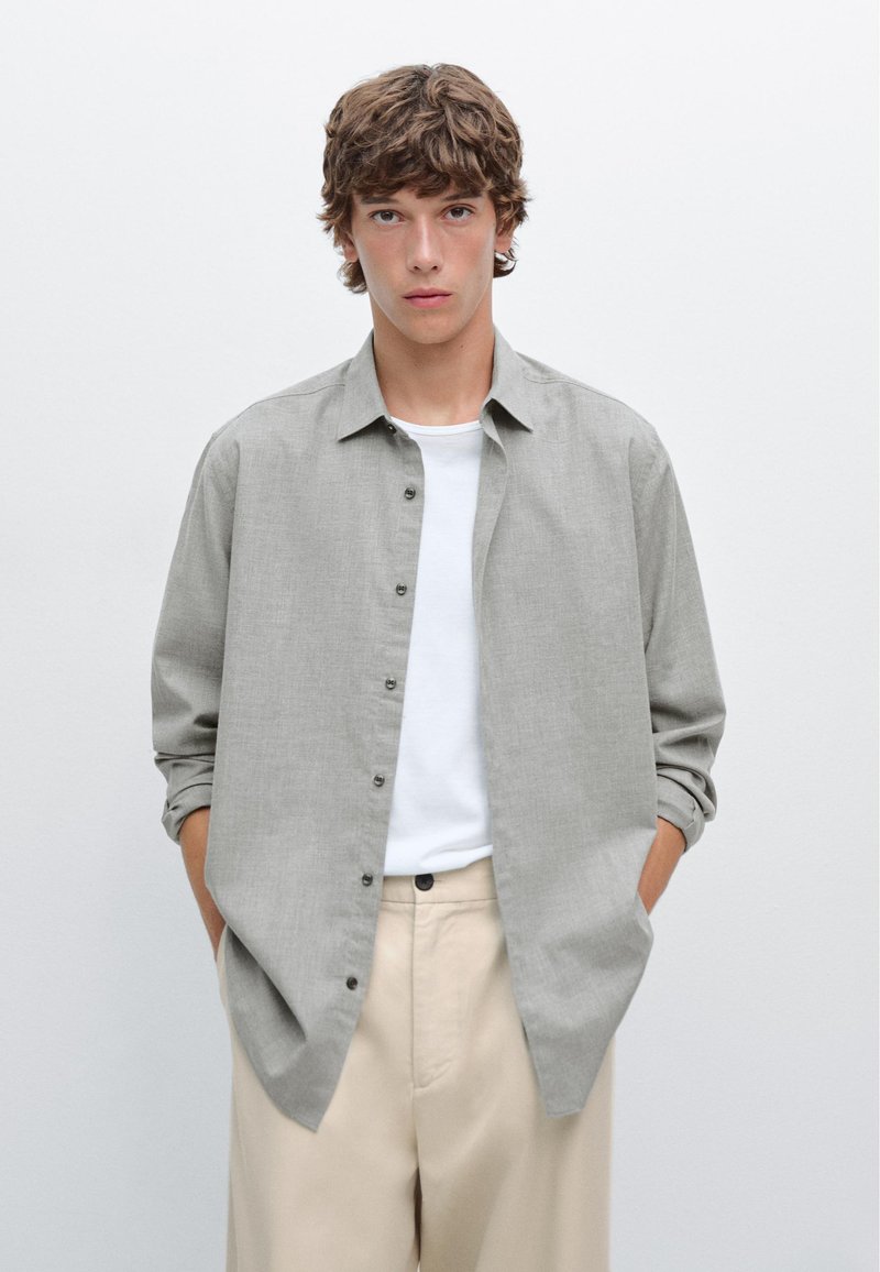 Lichtgrijze, overhemd met knopen en een zachte textuur, met een ontspannen pasvorm, gedragen over een wit T-shirt. Beige broek complementeert de outfit.