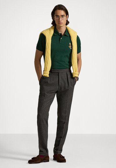 Camisa polo verde com detalhe bordado, combinada com calças cinzentas ajustadas. Colete amarelo de malha sobre os ombros. Sapatos mocassim castanhos.