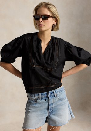 Femme portant des lunettes de soleil écaille de tortue, un chemisier noir à manches bouffantes avec des détails en dentelle, et un short en jean clair taille haute aux bords effilochés.