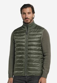 Grön pufferväst med quiltat mönster och hög krage, kombinerad med en stickad olivgrön tröja, som har dragkedja och ribbade mudd.