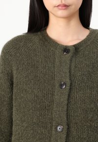 Cardigan verde măsliniu, fabricat dintr-un material moale și texturat, cu o cheeră rotundă, având patru butoane închise la culoare pe față.