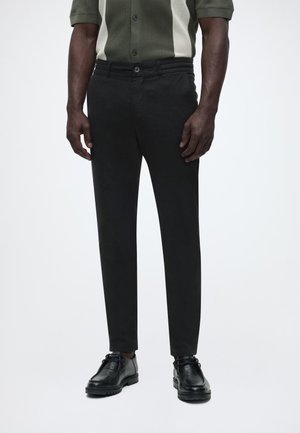 Mees, kes kannab musta kitsast pükst koos mustade pits-up kingade ja lühikeste varrukatega rohelise-valge nööpidega särgiga.