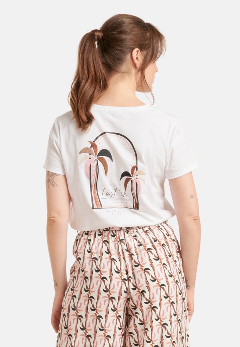 Witte katoenen t-shirt met een bedrukte design met palmbomen en de tekst "Verloren in de Woestijnparadijs." Korte mouwen, losse pasvorm.
