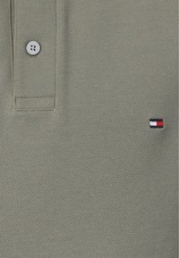 Ελιά πράσινη μπλούζα πόλο από υφαντό ύφασμα, με κολάρο δύο κουμπιών και μικρό κεντημένο λογότυπο σε ναυτικό μπλε, κόκκινο και λευκό.