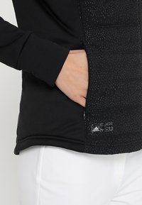 Veste de sport noire avec une texture matelassée d'un côté, un matériau lisse de l'autre, et une poche zippée ornée d'un logo.