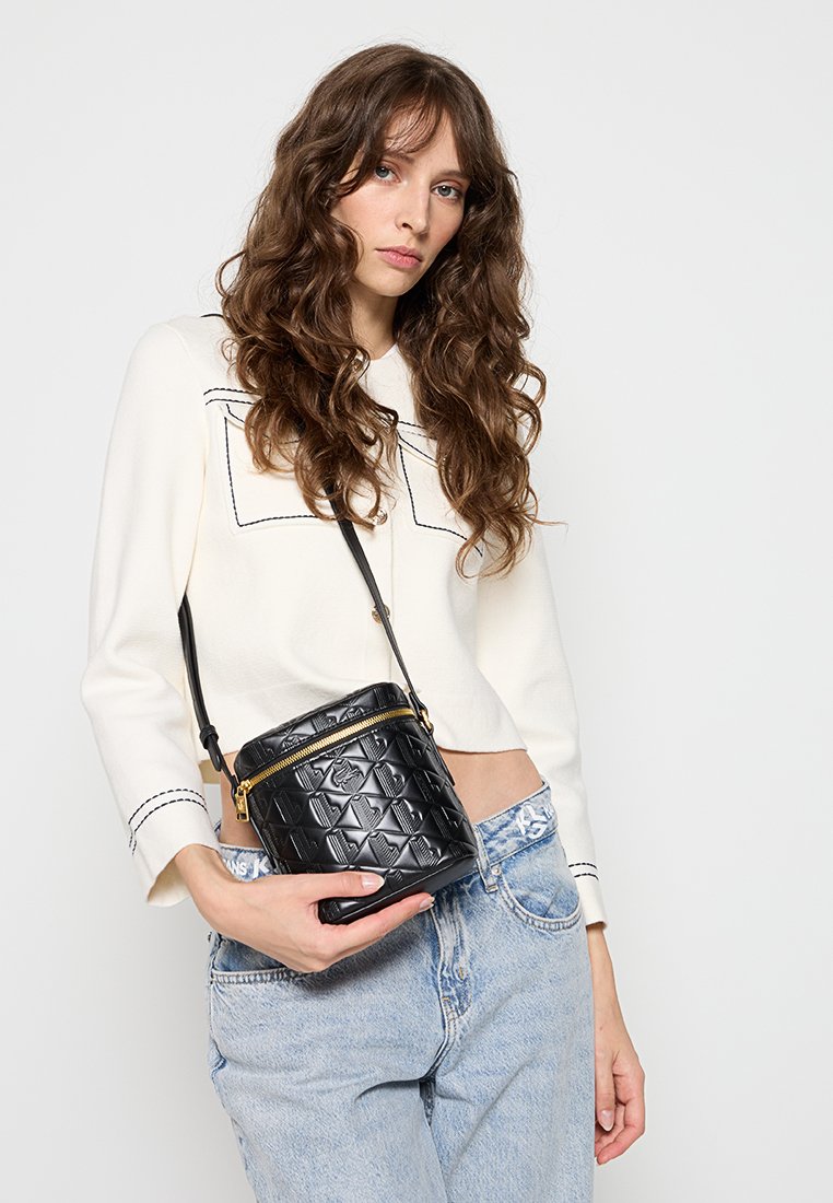 Sac en cuir noir en bandoulière avec motif embossé, fermeture éclair dorée et bandoulière réglable. Porté avec un haut blanc et un jean bleu clair.