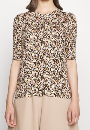 Vrouw in een korte mouw bloes met bloemen in roze, oranje en geel, gecombineerd met beige broeken.