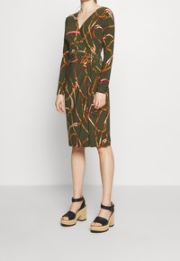 Kvinna i en knälång olivgrön omslagsklänning med röd och gul kedjeprint, kombinerad med svarta platåsandaler med ankelsnören.