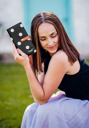 Black wallet with white polka dots, accented with a tan strap and gold hardware. Smooth texture, rectangular shape.