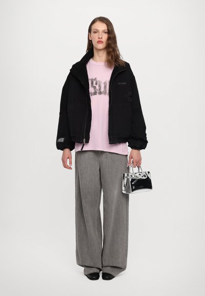Black jacket with a textured collar, pink graphic T-shirt, wide grey pants, and a small transparent handbag. Sleek black shoes complete the look.