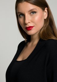 Cardigan noir avec un décolleté en V profond. Texture lisse, porté sur un top noir sans manches. Le modèle a de longs cheveux raides et porte du rouge à lèvres rouge.