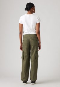 Cargo-Hosen aus olivgrünem Baumwollstoff mit elastischen Bündchen, ausgestattet mit Taschen an den Seiten und auf der Rückseite. Kombiniert mit einem einfarbigen weißen T-Shirt mit kurzen Ärmeln.