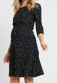 Kobieta w ciąży ubrana w czarną sukienkę w białe i niebieskie kształty serc, z rękawami o długości trzech ćwiartek i cienkim paskiem w talii.