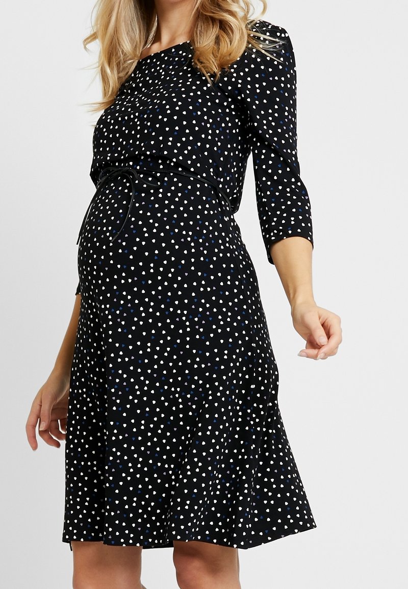 Kobieta w ciąży ubrana w czarną sukienkę w białe i niebieskie kształty serc, z rękawami o długości trzech ćwiartek i cienkim paskiem w talii.