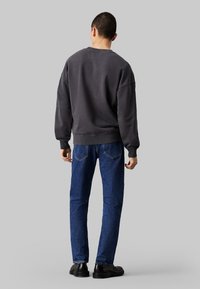 Hombre de espaldas vistiendo un sudadera oversized gris oscuro, jeans azules y zapatos negros frente a un fondo gris plano.