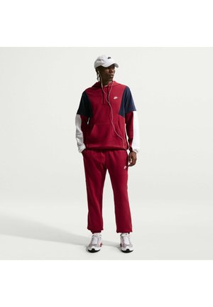 Mężczyzna ubrany w czerwony, granatowy i biały dres marki Nike, białe trampki, biała czapka i słuchawki, stojący na tle jednolitego jasnego tła.