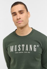 Grön långärmad t-shirt i mjukt tyg med rund halsringning, med en stor vit "MUSTANG"-logotyp och mindre text under.