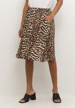 Kvinde iført en knælængde nederdel med leopardprint, hvid top og sorte hælede mule sandaler med åben tå, stående mod en ensfarvet lys væg.