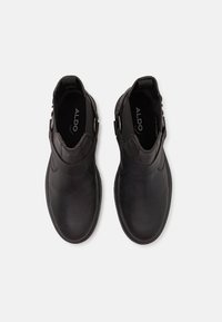 Botas de cuero negras hasta el tobillo con una textura suave, que presentan un detalle de hebilla lateral y punta redonda, situadas planas sobre un fondo blanco.