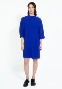 Modra prevelika obleka z visokim ovratnikom in kratkimi rokavi, izdelana iz mehkega materiala. V kombinaciji s črnimi loaferji, stoji proti beli ozadju.