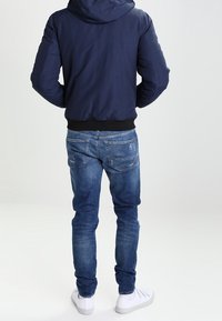 Mann trägt marineblaue Kapuzenjacke, blaue Jeans und weiße Sneakers, steht mit den Händen in den Taschen und schaut auf einfachem Hintergrund nach hinten.