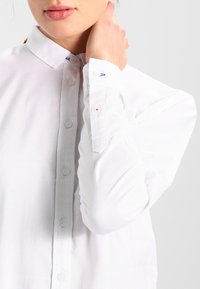 Camisa blanca de manga larga con botones y cuello clásico. Presenta una tela texturizada y un pequeño detalle del logo cerca del escote.