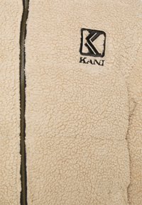 Beige fleecejakke med en teksturert overflate, med svart glidelås og brodert "KANI"-logo på brystet.