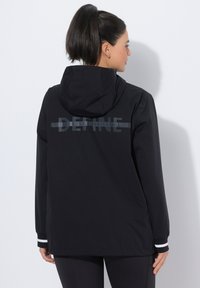 Czarna kurtka z kapturem o teksturowanej powierzchni, z graficznym wzorem na plecach, elastycznymi mankietami z białymi paskami i luźnym krojem.