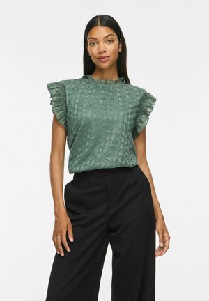 Femme portant un chemisier vert texturé à manches courtes avec des détails à volants, associé à un pantalon noir taille haute à jambes larges.