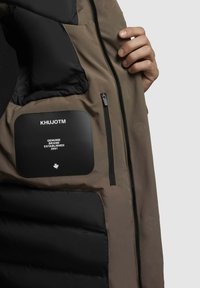 Interior de chaqueta acolchada en marrón y negro. Presenta una etiqueta negra con "KHUJOTM", bolsillos y una cremallera en una tela texturizada.