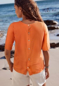 Oranssi pellavapusero, jossa on lyhyet hihat, avonainen selkä ja neljä nappia. Päällä valkoiset shortsit, taustalla rantamaisema.