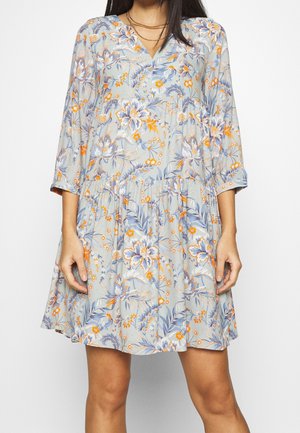 Femme portant une robe fleurie gris clair avec des fleurs orange et bleues, manches trois-quarts, et un décolleté en V.